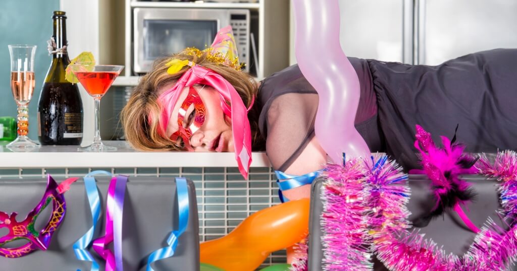 Woman in party accessories slumped across a kitchen counter surrounded by cocktails, champagne, balloons and streamers.