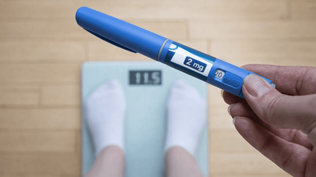 Close-up of a weight loss injection pen held above bathroom scales, illustrating the reality of GLP-1 weight loss jabs and their risks beyond the numbers on the scale.